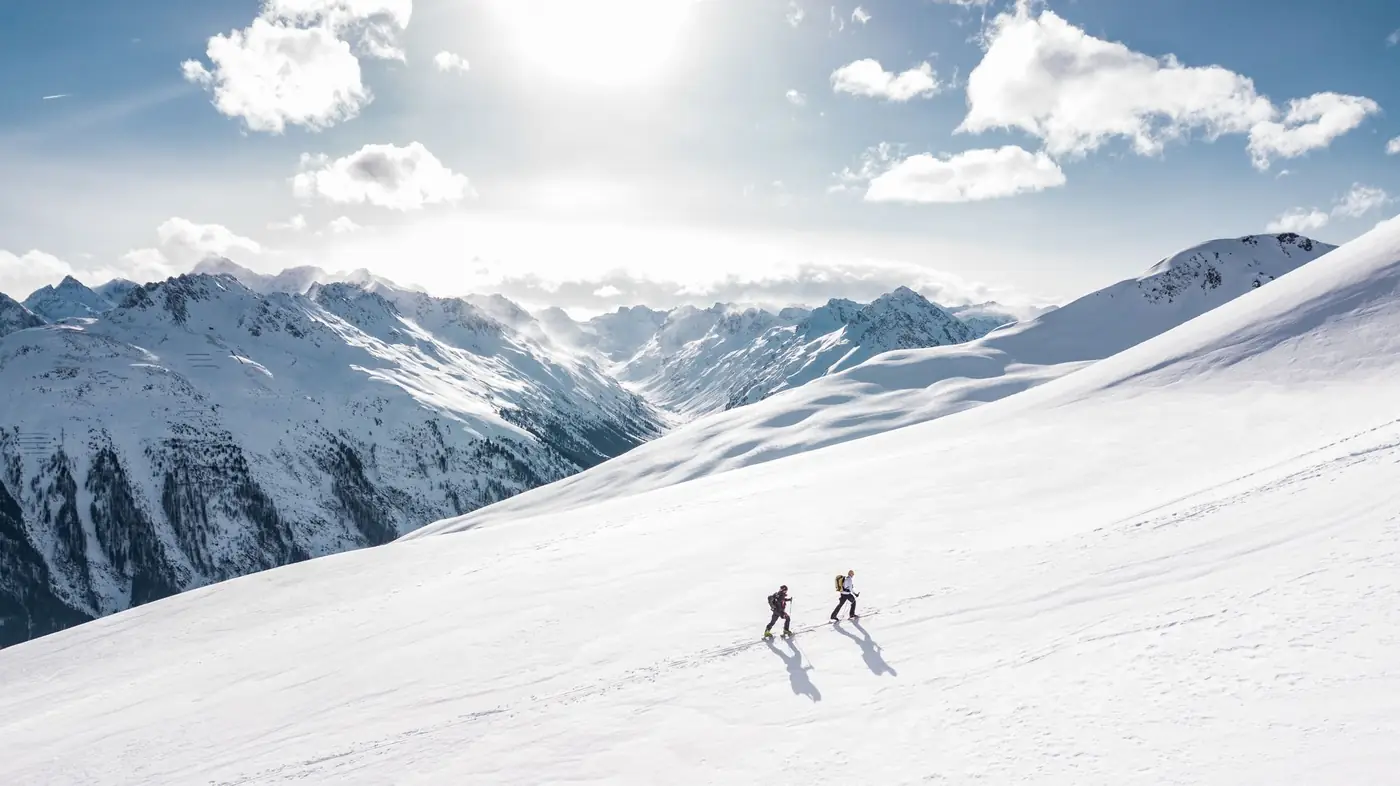 Twee wintersporters lopen met ski's over een zonnige besneeuwde berghelling