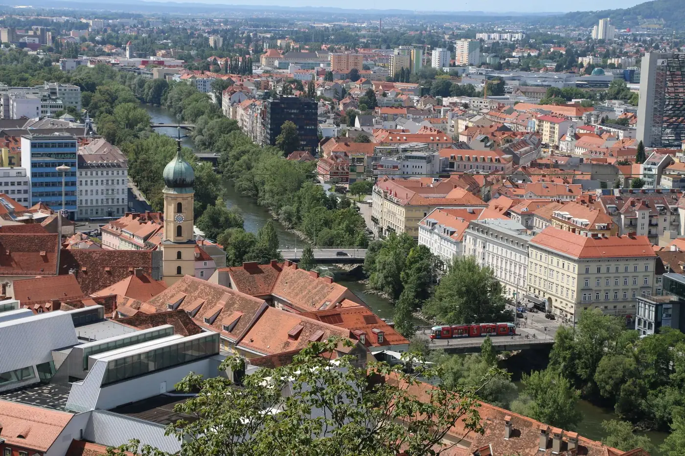 A scenic aerial view of Graz, Austria featuring red rooftops, the Mur River, and lush greenery.