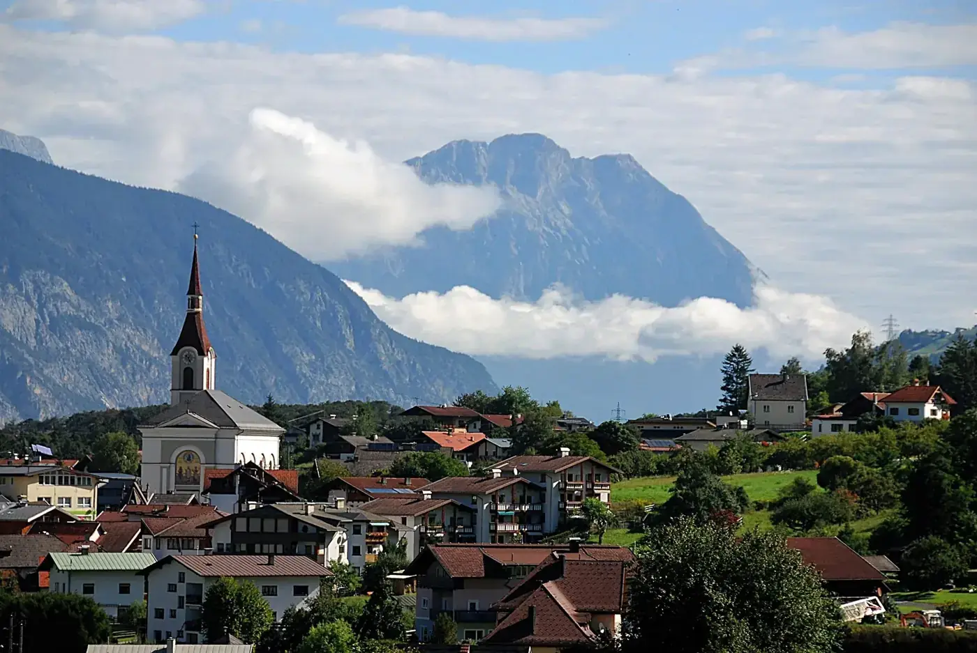 Scenic view of a picturesque Alpine village with a church, nestled against a backdrop of majestic mountains under a blue sky.