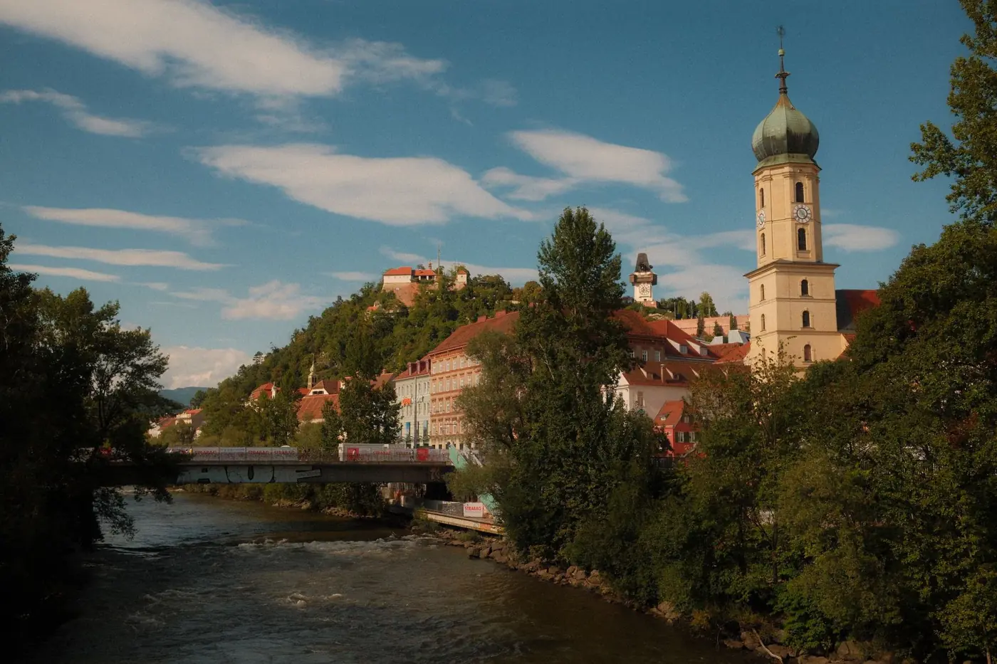 Picturesque landscape of Graz, Austria featuring the historic Uhrturm and cascading river under a bright sky.