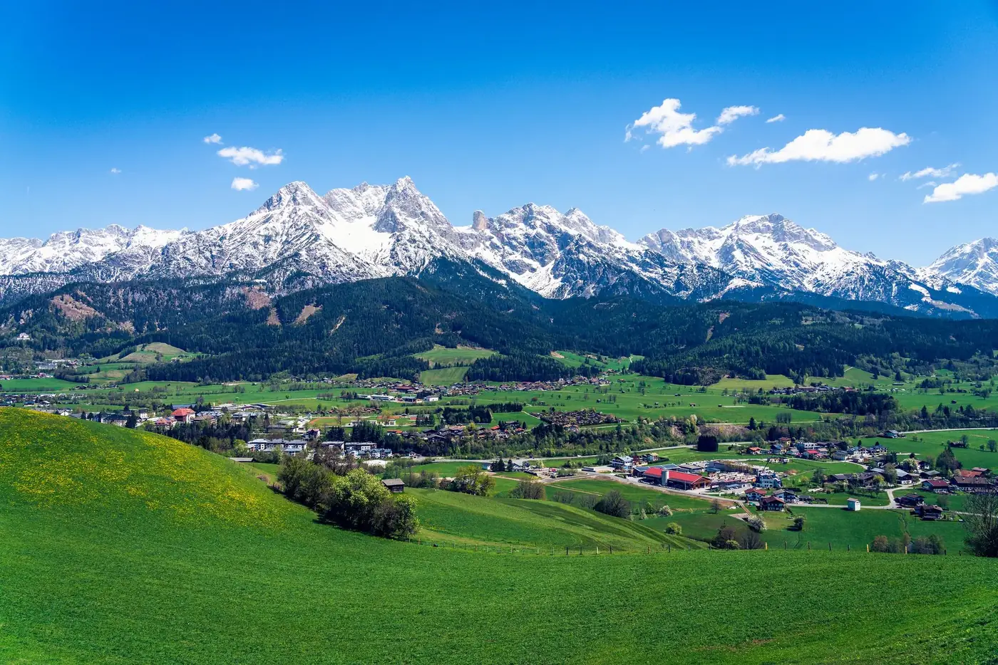 Panoramic view of snow-capped mountains and lush green fields under a clear blue sky.