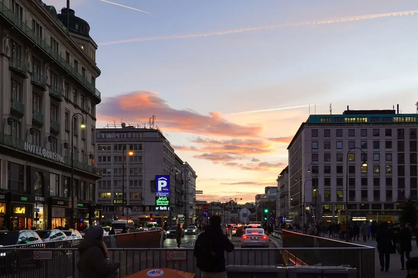 Busy Vienna street at sunset with people and cars under a vibrant sky.