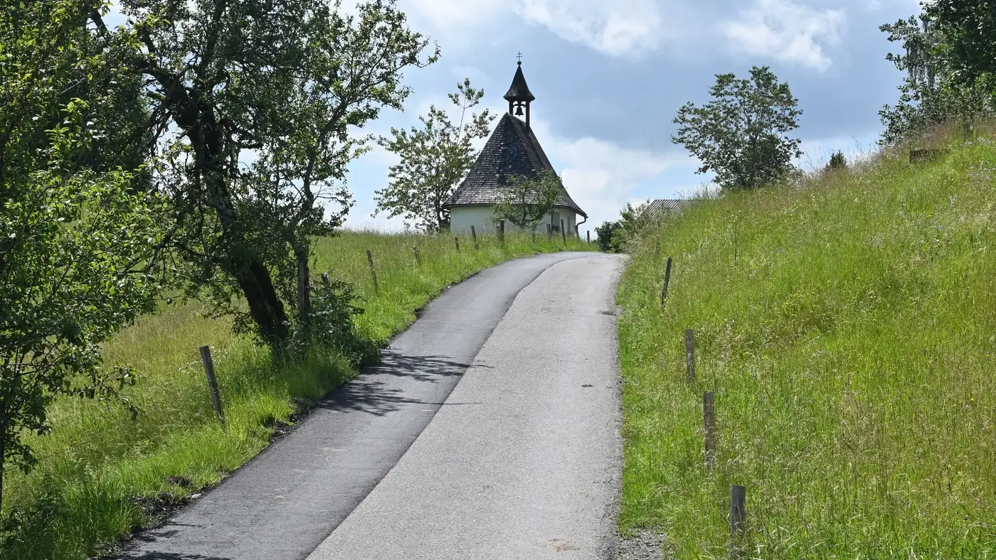 Serene view of a chapel along a rural road in the lush summer landscape of Hittisau, Austria.