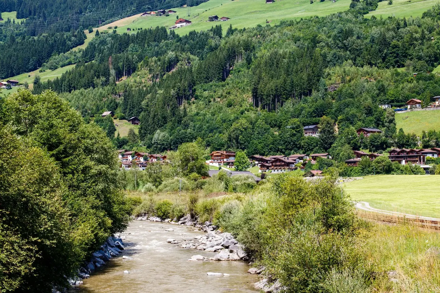 Charming alpine village nestled by a serene river and lush green hills under a bright sky.