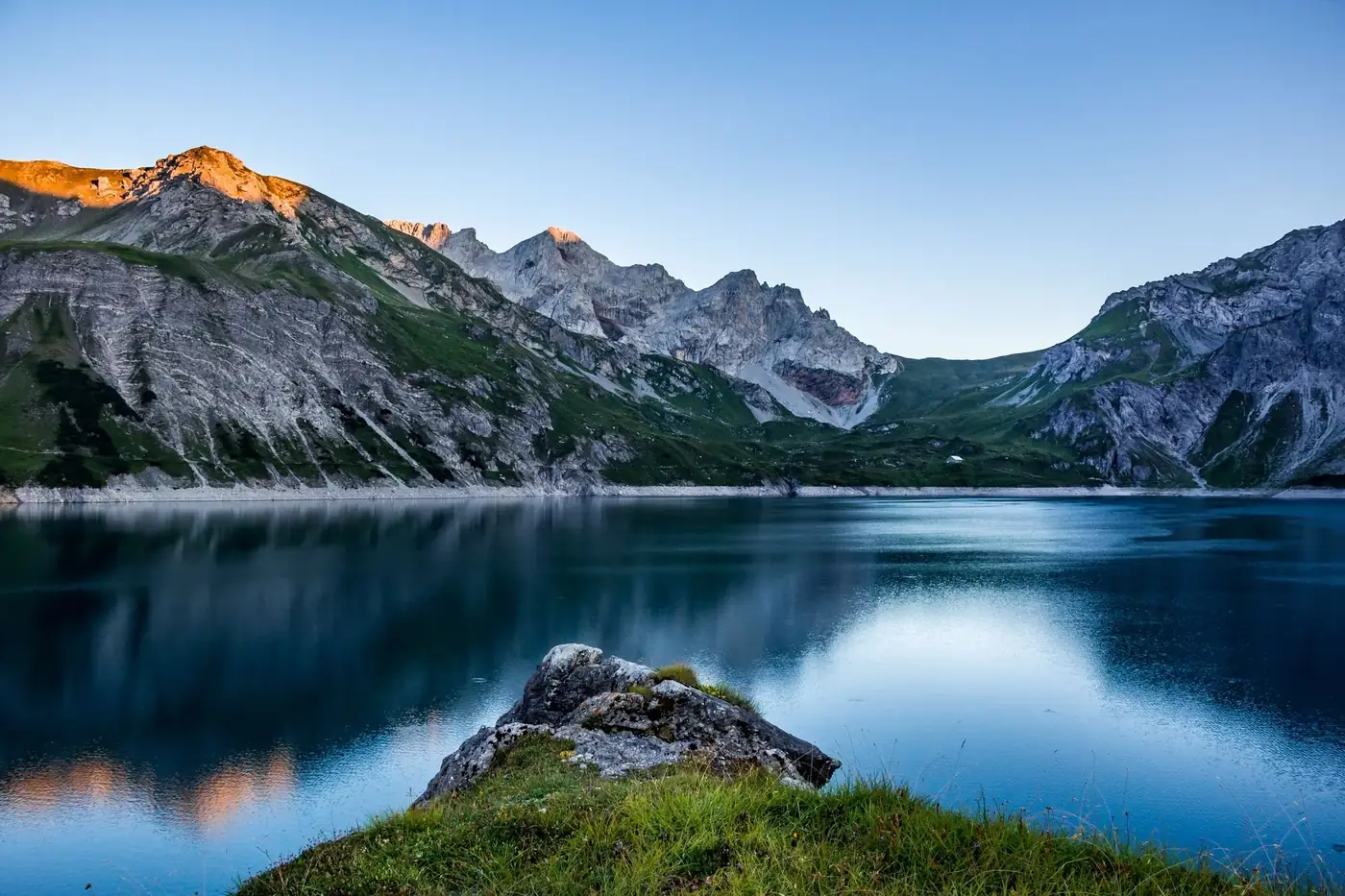 Serene Austrian alpine lake surrounded by majestic mountains and lush greenery at sunrise.