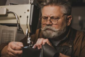 senior, bearded cobbler sewing leather on sewing machine